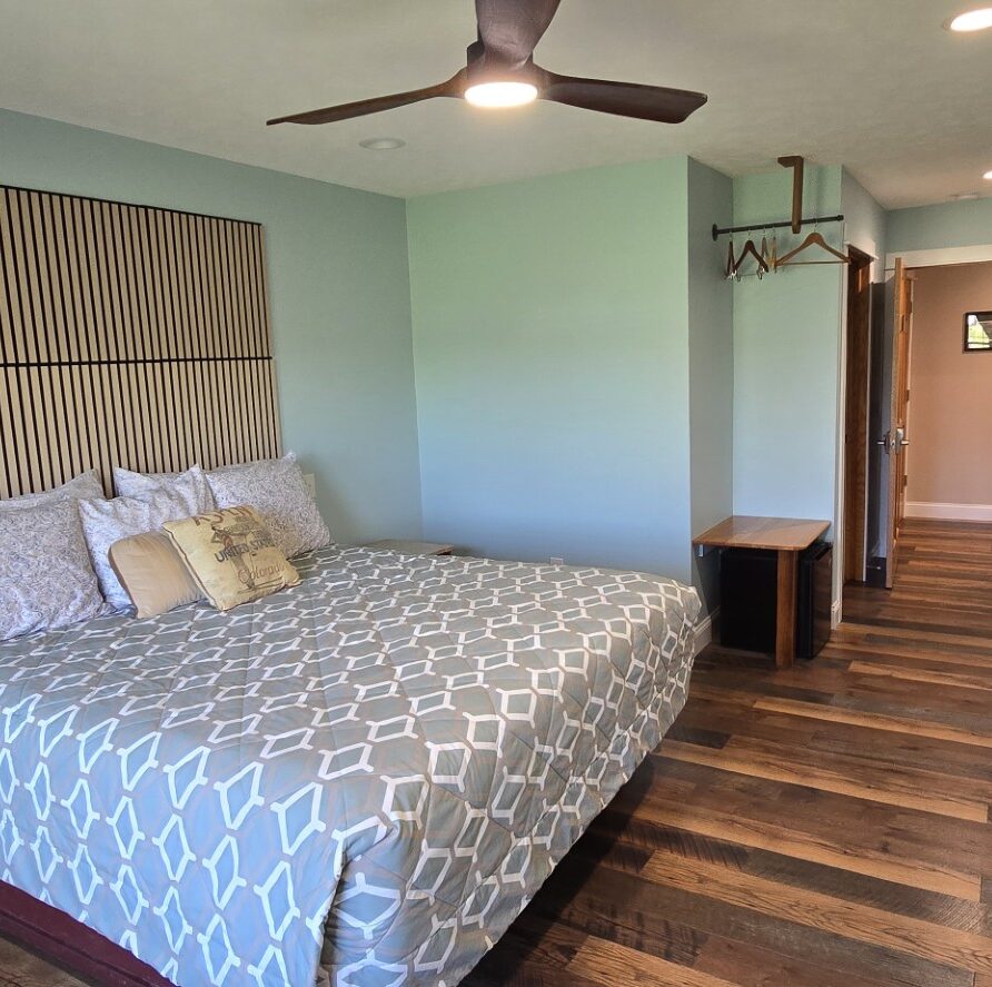 20260521_174658 - London Guest House King-size bed with a geometric gray-blue comforter and multiple pillows, a tall striped wooden headboard, and teal walls in a modern hotel room. A ceiling fan above, wood-laminate floor, and small desks flank the bed. Opposite wall has a wall-mounted TV. A hallway leads to the door.
