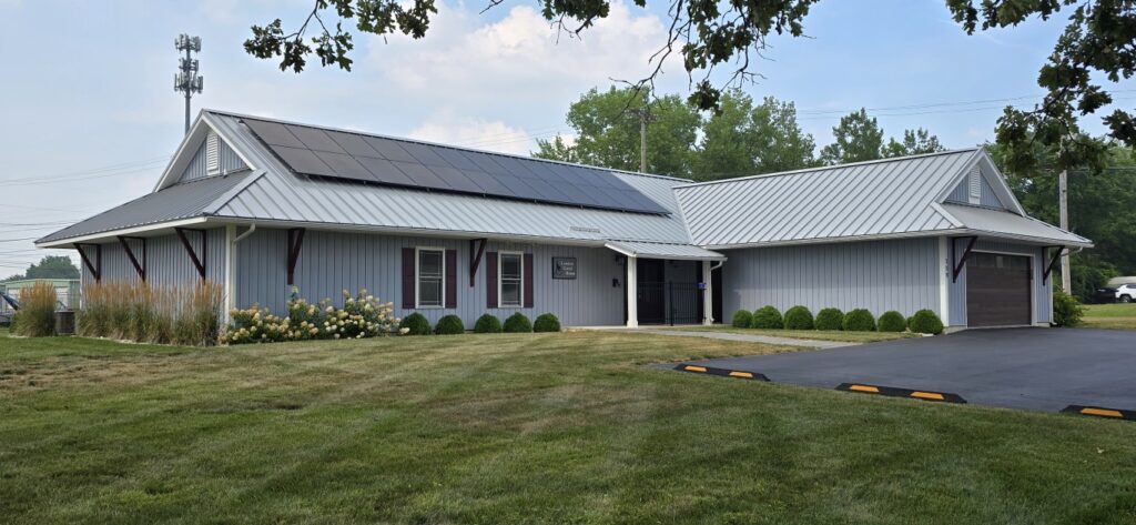 A light-blue metal-roofed building with solar panels, connected wings, and a driveway leading to a closed garage door in a grassy setting.
