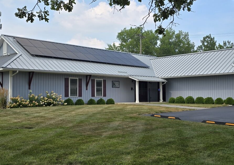 A light-blue metal-roofed building with solar panels, connected wings, and a driveway leading to a closed garage door in a grassy setting.