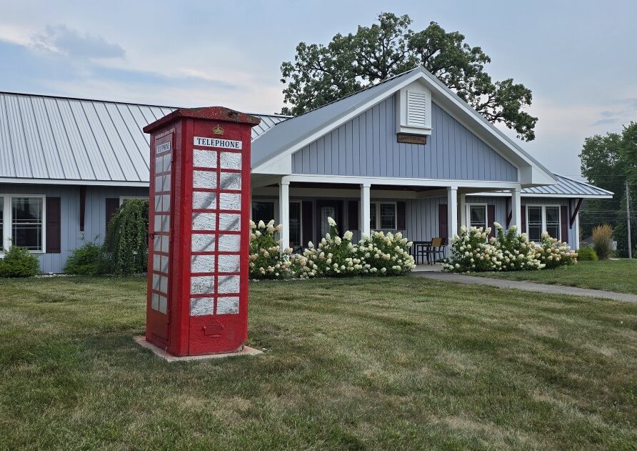 Red British-style telephone booth on a grassy front yard in front of a blue house with a covered porch and white roses.