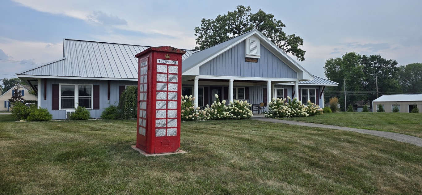 20260717_161753 - London Guest House Red British-style telephone booth on a grassy front yard in front of a blue house with a covered porch and white roses.