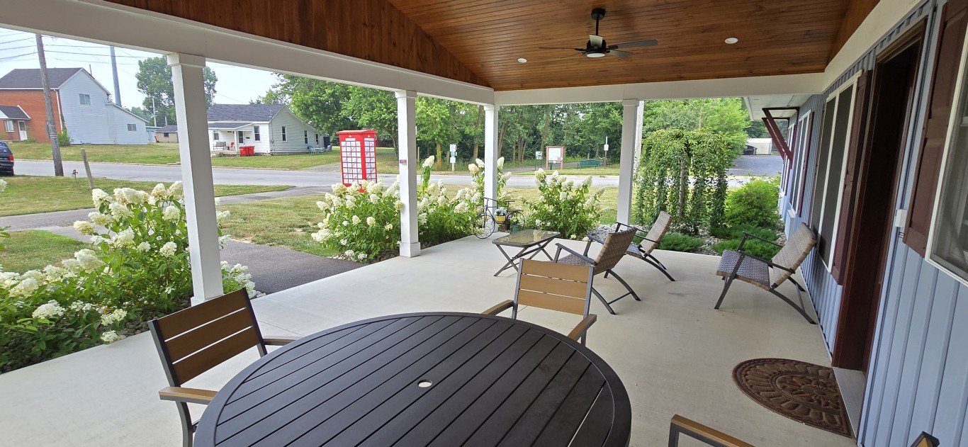 20260717_161922 - London Guest House Covered porch with a round wooden table and several lounge chairs, white hydrangea bushes along the edge, and a residential street in the background.