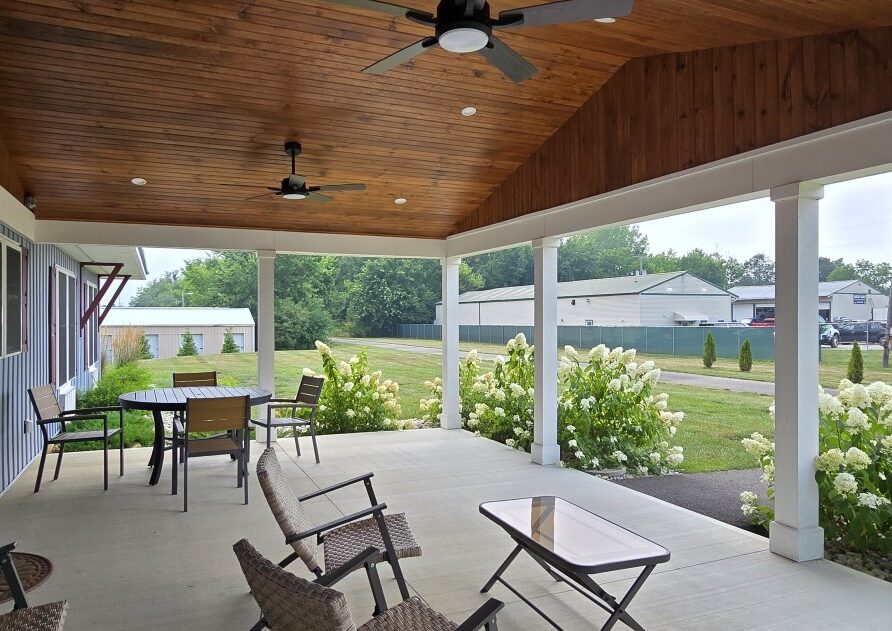 Covered patio with a wooden ceiling and ceiling fans, outdoor table and chairs, and white hydrangeas along the edge overlooking a lawn.