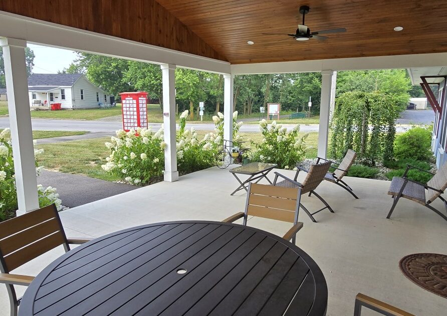 Covered porch with a round black table and wooden chairs, hydrangea bushes along a walkway in a quiet neighborhood.