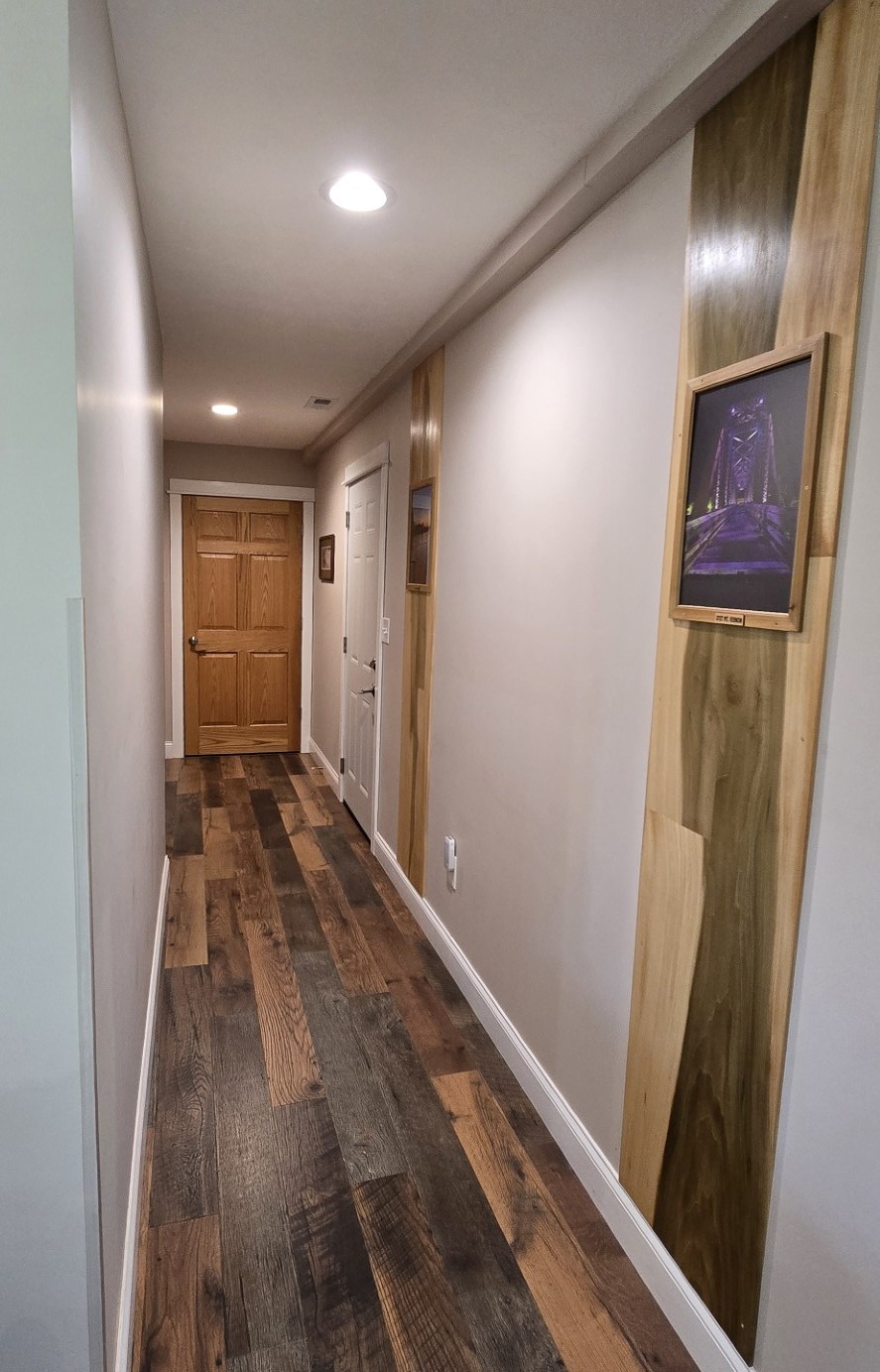 20260521_175022edit - London Guest House Long residential hallway with wood plank flooring, beige walls, and a wooden door at the end; framed artwork mounted on a wood-paneled wall to the right.