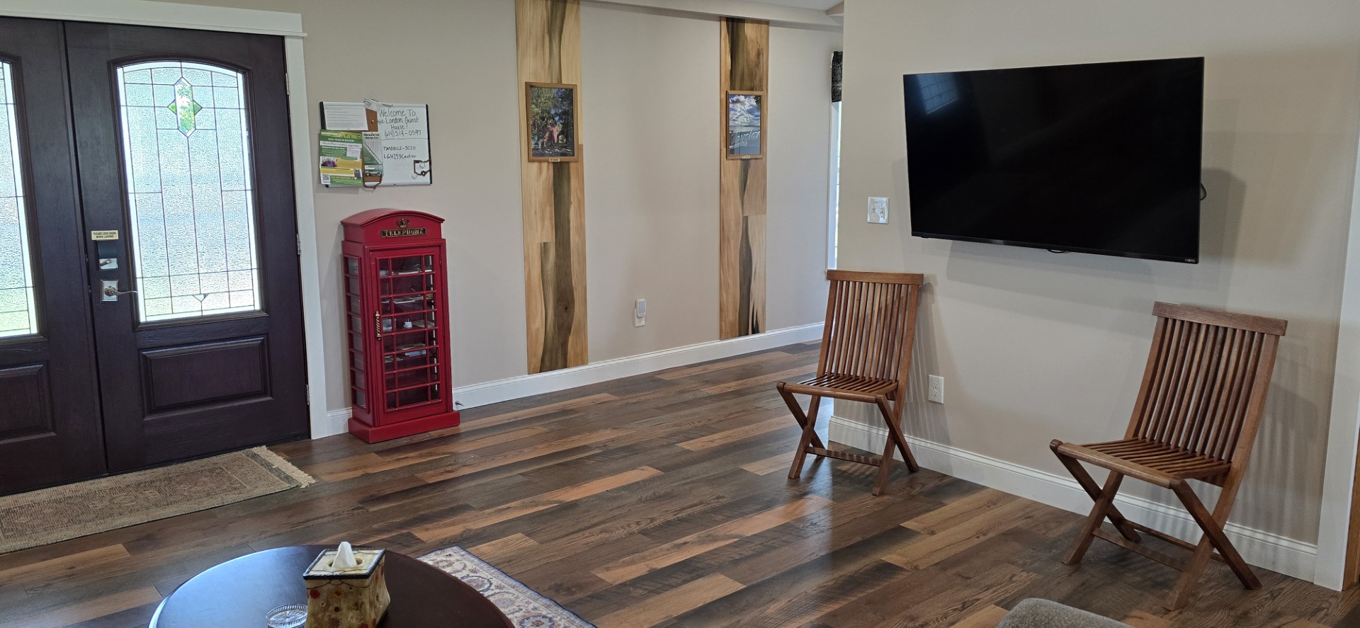 20260521_175135 - London Guest House Cozy lobby corner with dark wooden front doors, a red vintage telephone booth cabinet, wall art panels, a mounted flat-screen TV, and two wooden chairs.